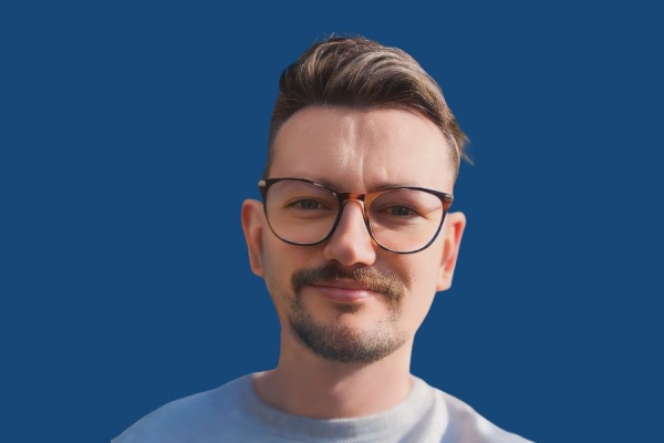 A young white man with a blond quiff, a beard and moustache wearing glasses on a blue background